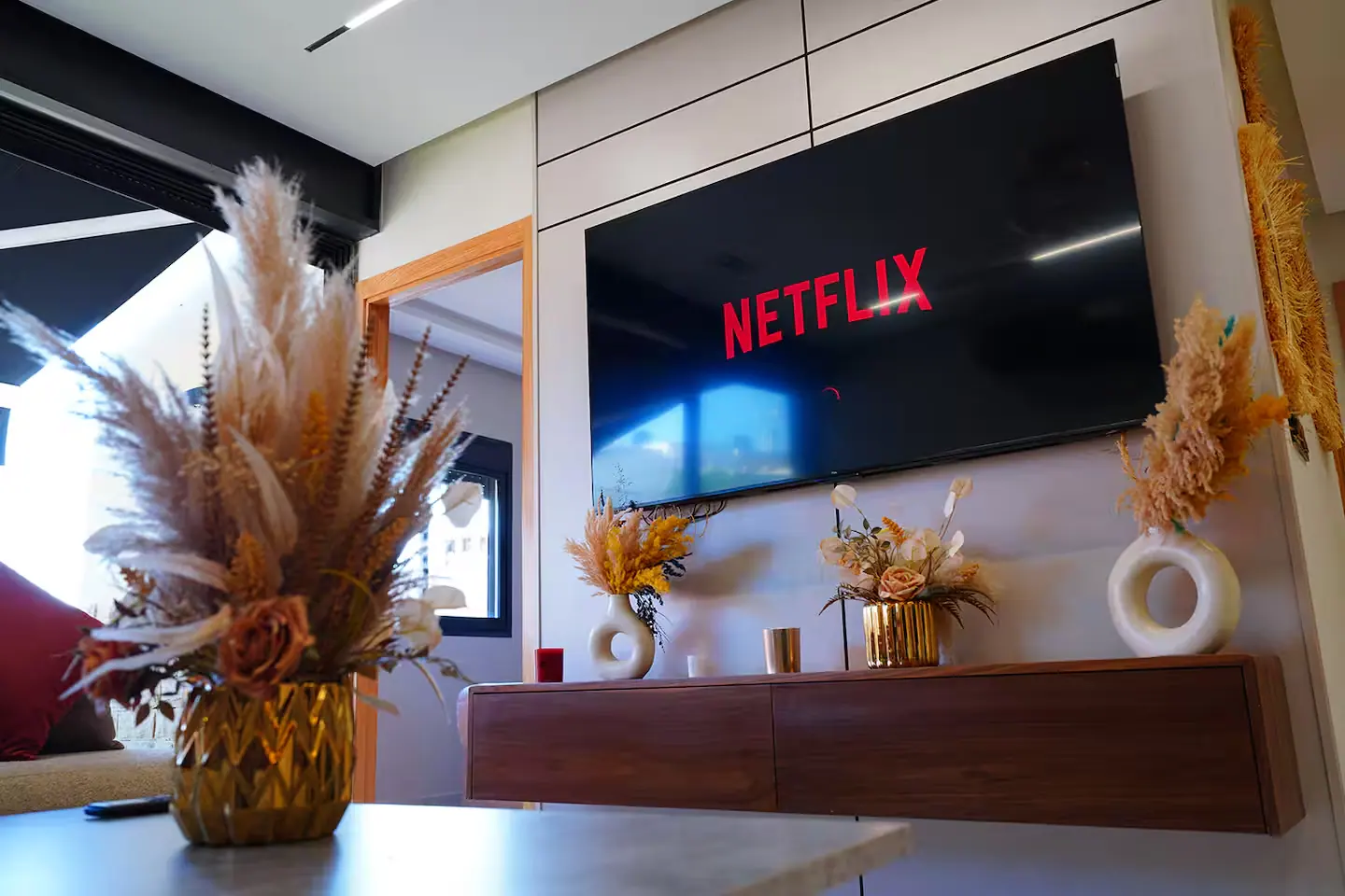 Coin salon d'un logement à Rabat avec meuble télé en bois, bouquets de fleurs séchées et grande baie ouverte sur la terrasse