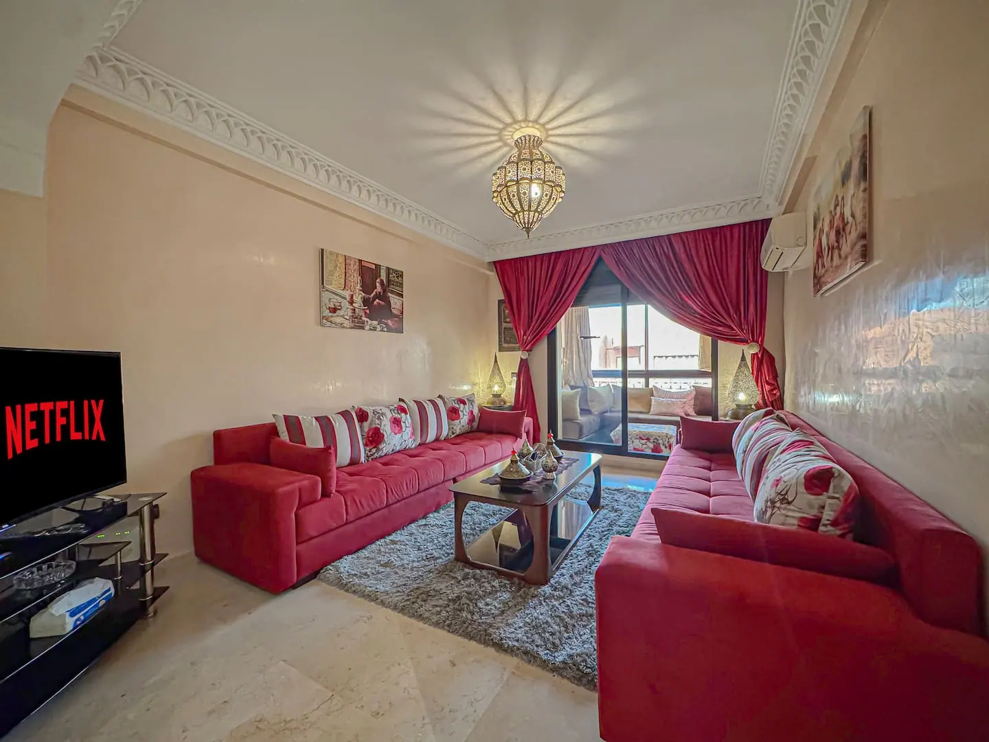Salon marocain avec banquettes rouges, lustre en laiton ciselé et grande baie vitrée dans un appartement du quartier Guéliz à Marrakech
