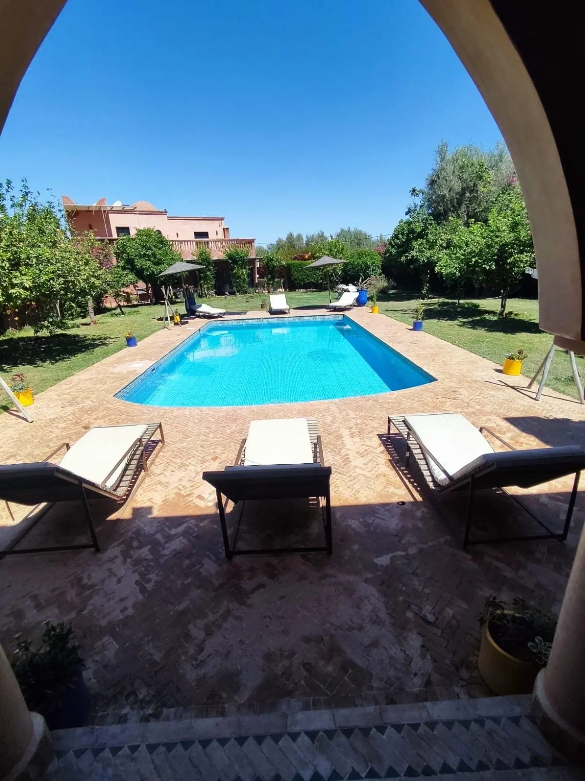 Piscine rectangulaire bordée de transats et de dalles en terre cuite, vue depuis une arcade d'une villa de la région de Marrakech, avec jardin arboré et maison ocre en arrière-plan