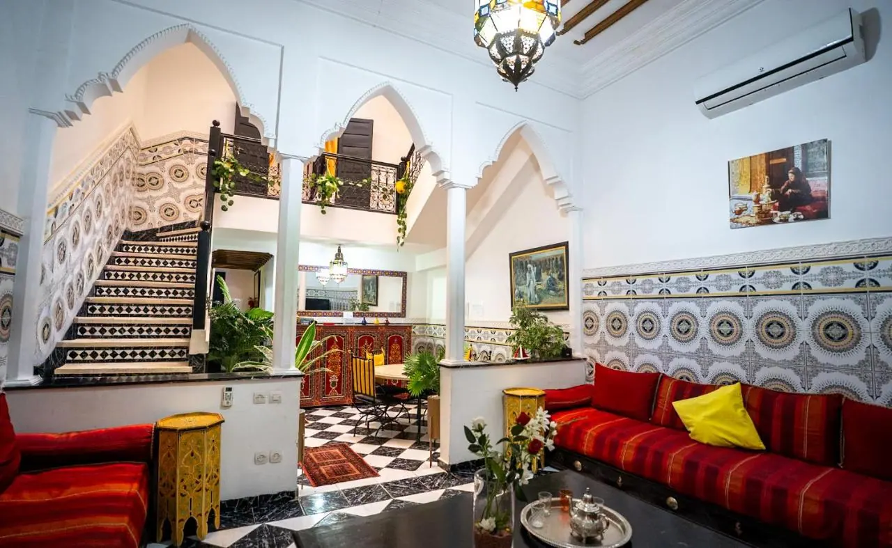 Grand salon d'un riad marocain avec banquettes rouges, zellige noir et blanc, arcades sculptées et escalier en mosaïque, à Marrakech