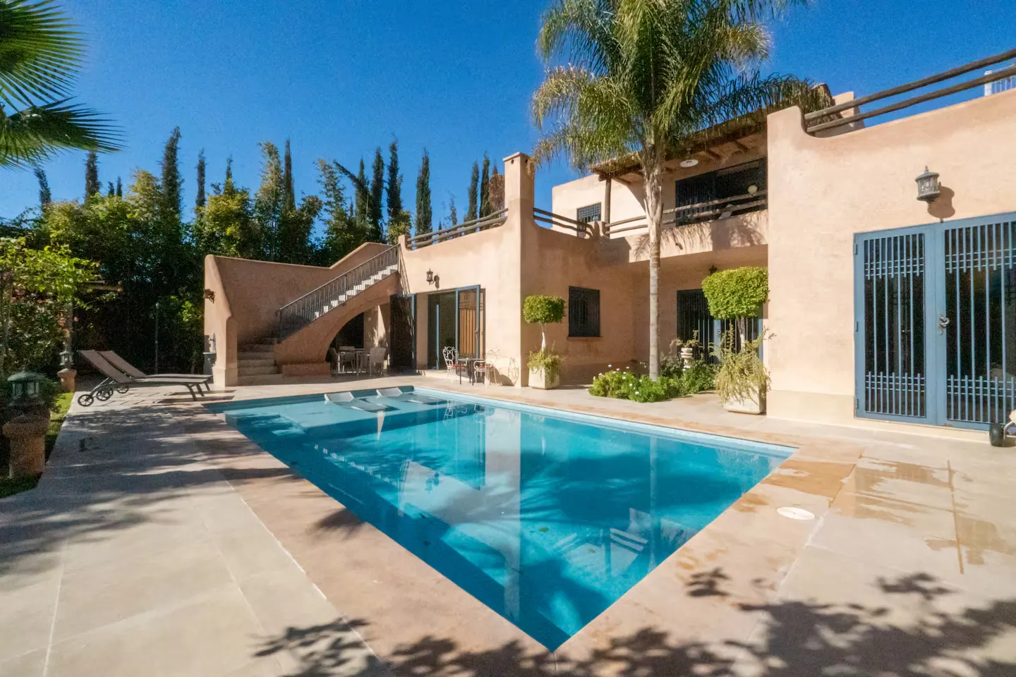Piscine privée d'une villa marrakchie entourée de transats et d'un salon extérieur couvert, avec vue sur le jardin